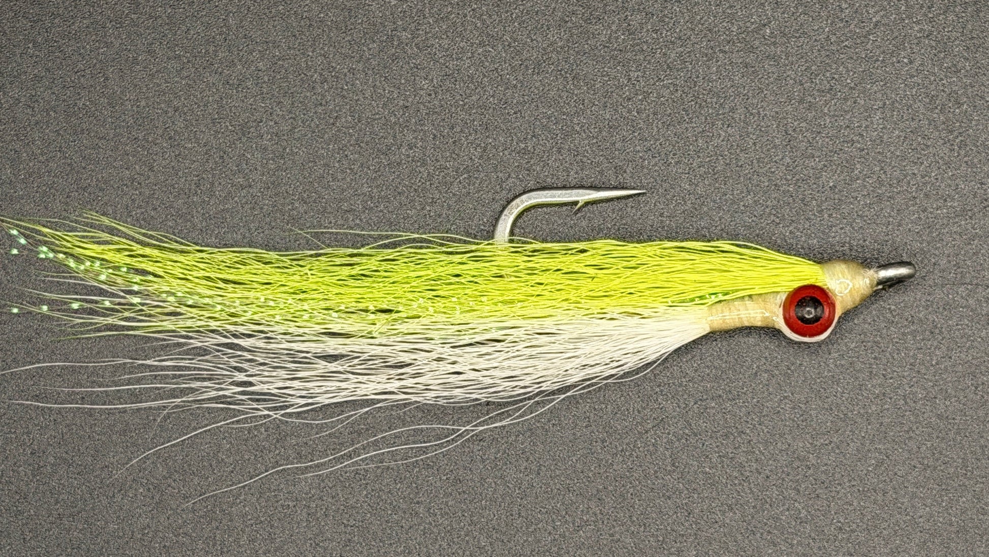 A fly fishing lure with a bright chartreuse yellow-green body, a white skirt, and a red-and-black eye, shown on a dark gray textured background.